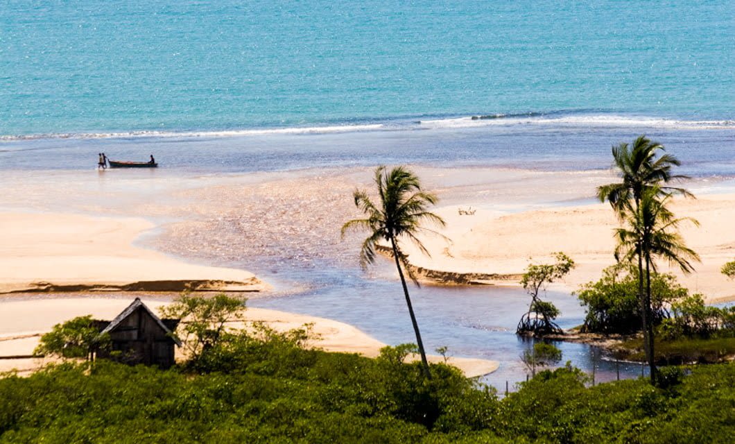 A serene tropical beach with turquoise water, white sand, palm trees, green shrubs, and a small wooden hut awaits you—a perfect retreat after an exciting Brazil Golf Tour. Nearby, a boat with two people rests gently along the shoreline.