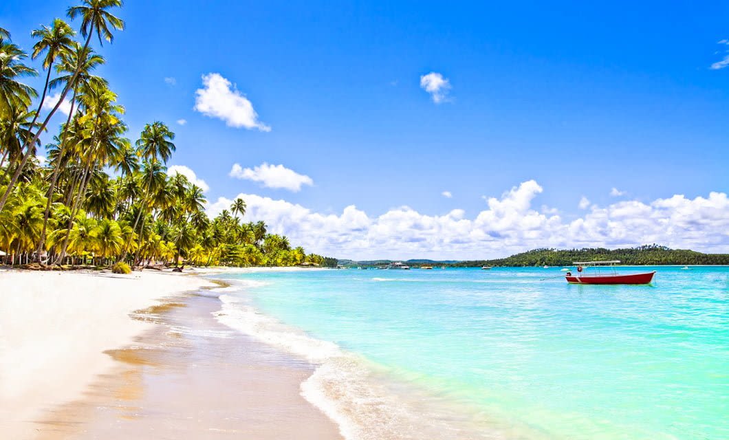 Sunny tropical beach on Marajó Island with white sand, turquoise water, and tall palm trees lining the shore near Salvaterra. A red and white boat floats on the calm sea under a bright blue sky with scattered clouds.