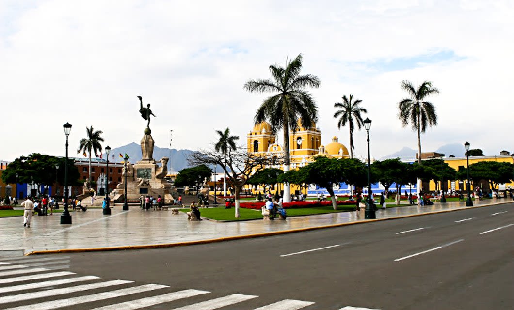A city square in Trujillo with palm trees, a yellow cathedral in the background, a statue in the center, people walking, and a crosswalk in the foreground under a partly cloudy sky.