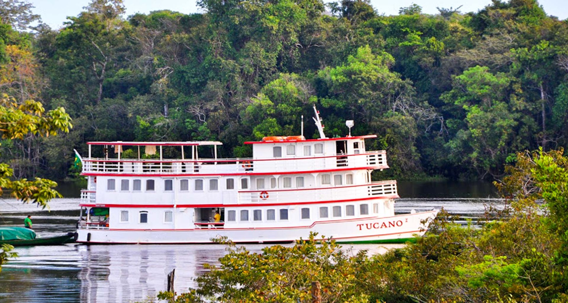 The Tucano, a white and red riverboat, sails on a calm river surrounded by dense green forest. Perfect for a Brazil Amazon River Cruise, it features multiple decks and windows, with trees beautifully reflected in the water.