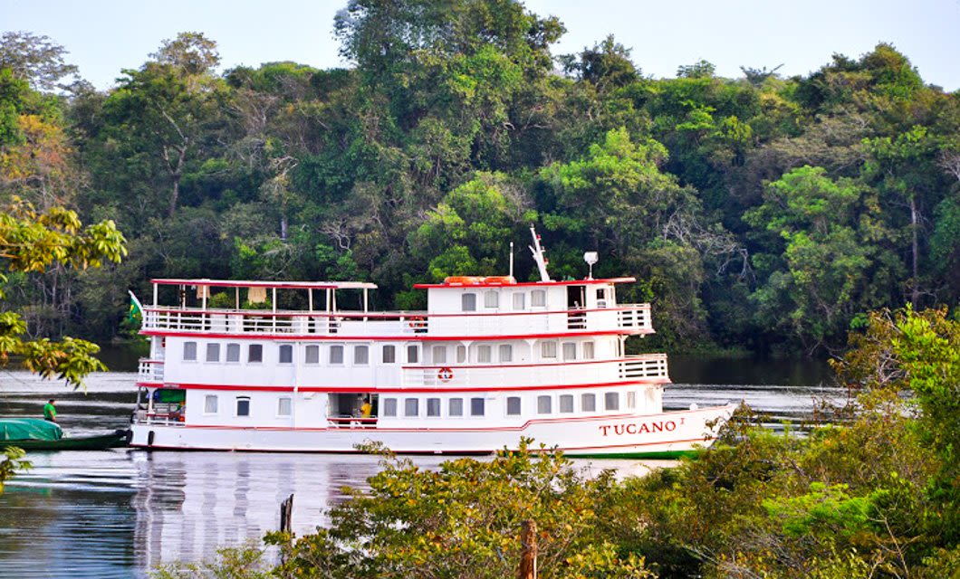 A white and red riverboat named Tucano floats on calm water, surrounded by lush, green South American forest under a clear sky. The boat, perfect for a Deluxe Tour, features multiple decks with railings and windows.