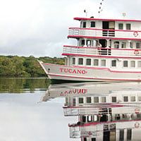 A white and red riverboat named Tucano is reflected on calm water, with green trees and a cloudy sky in the background, capturing the serene beauty of a Brazil Amazon River Cruise.