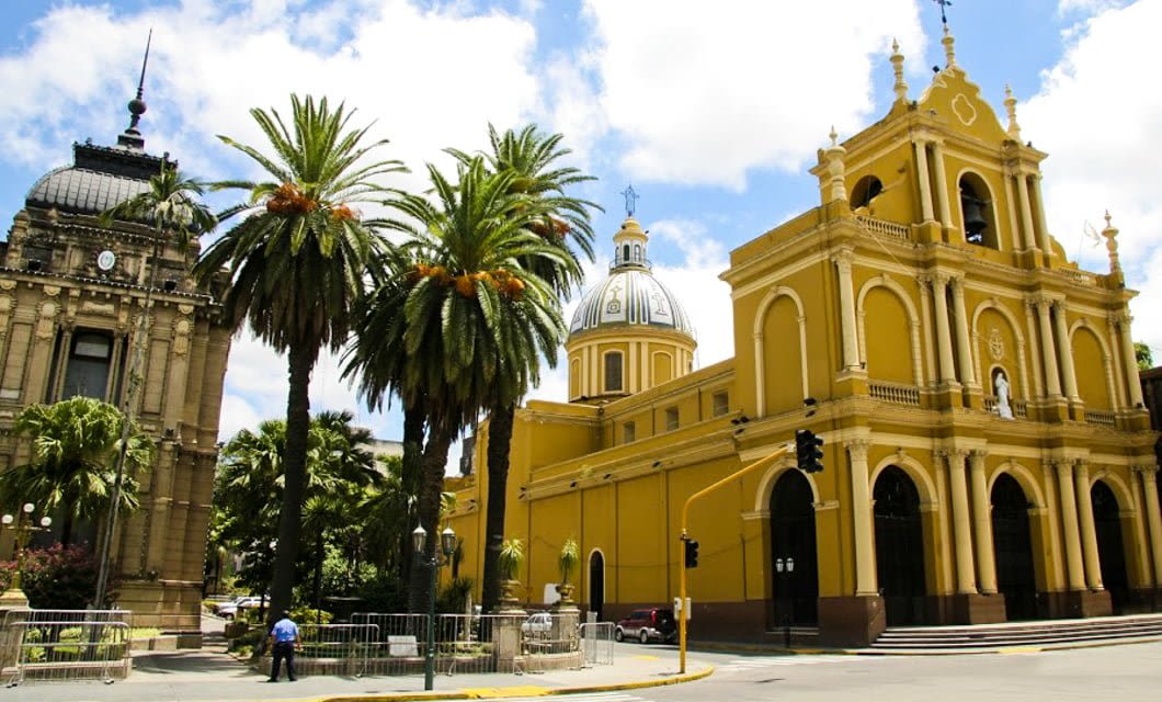 A sunny day in Northern Argentina with a bright yellow cathedral featuring domes and arches, palm trees nearby, and an ornate historic building to the left; a person in blue stands by a fence, capturing the region's wonders.