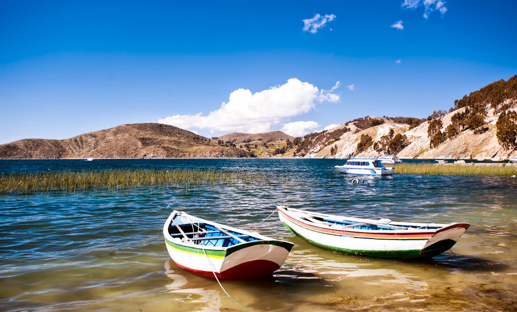 Two colorful rowboats float on a clear, blue lake in the Andes, surrounded by grassy shores and rolling hills under a bright, sunny sky with scattered clouds. A larger boat is seen in the background near the shore.