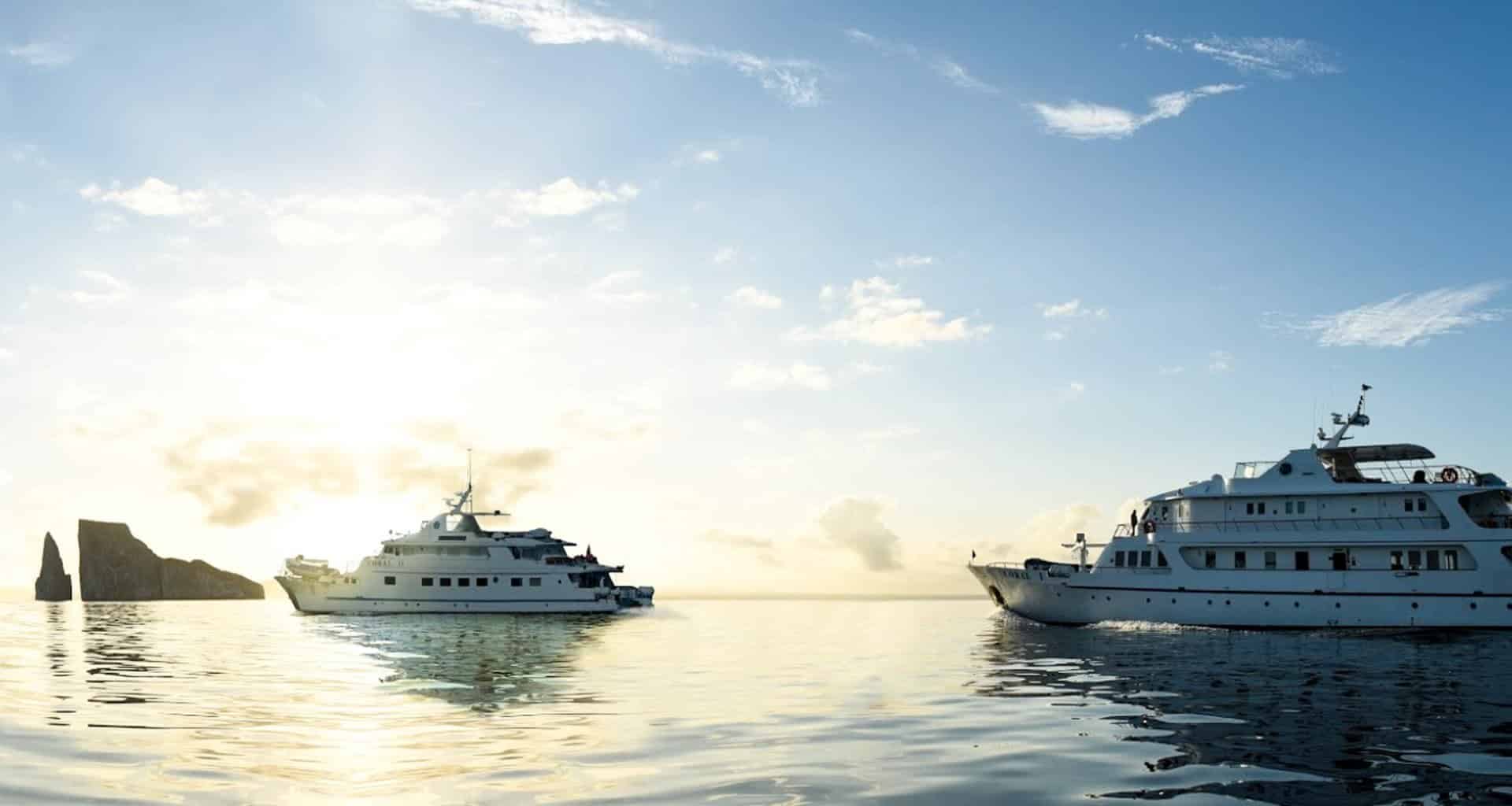 Two white yachts float on calm water at sunrise or sunset, the sun low on the horizon and a rocky island visible in the background. Experience serene beauty with Coral Cruises in the Galapagos under a partly cloudy sky.