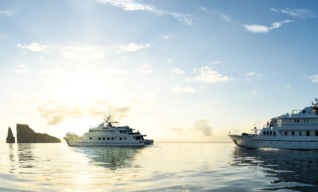 Two white yachts from Coral Cruises float on calm water at sunrise, with a rocky Galapagos island formation visible in the distance beneath a partly cloudy sky.