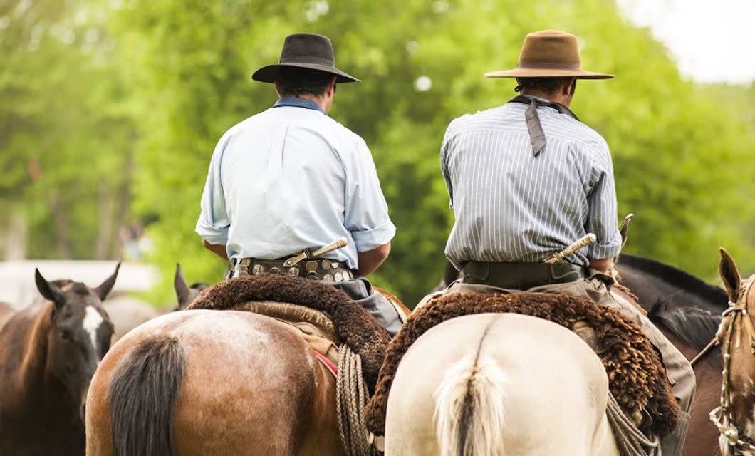 Two people wearing hats and traditional clothing ride horses side by side, seen from behind, with green trees in the background—evoking the spirit of a Buenos Aires countryside adventure.