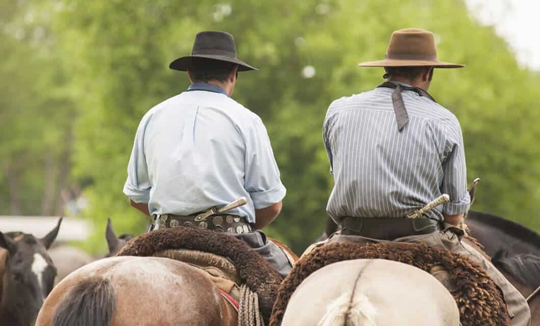 Two people in wide-brimmed hats and traditional clothing ride horses side by side through the lush landscape of Wild Argentina, green trees stretching into the distance behind them.