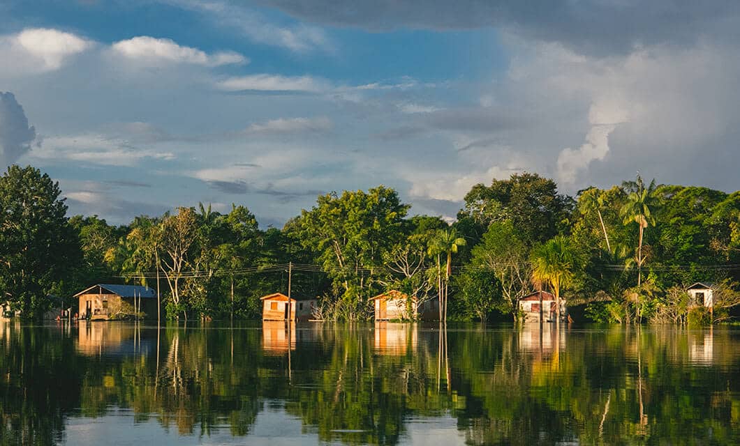 Small wooden houses at Uakari Lodge line the edge of a calm river, surrounded by lush green trees and reflecting on the water under a partly cloudy sky.