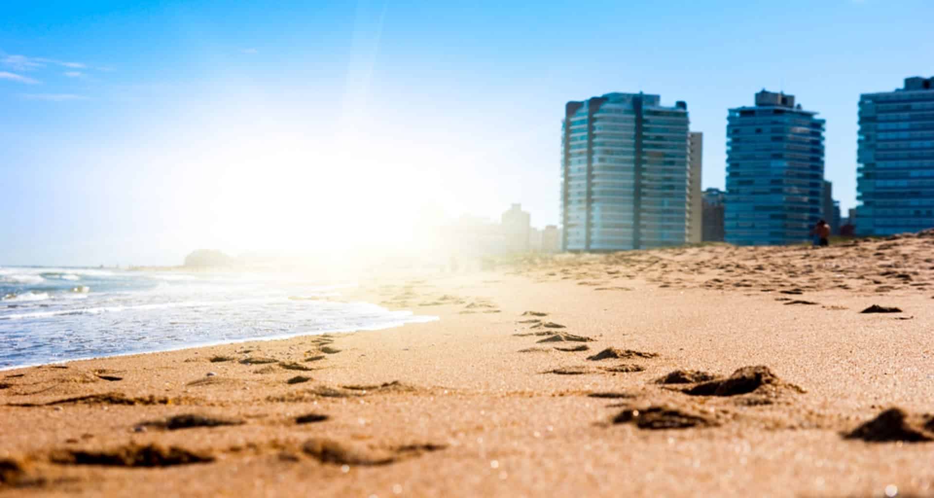 Footprints in the sand lead along a sunny beach in Punta del Este towards the ocean, with tall modern buildings and a bright blue sky in the background. The sun shines brightly, creating a warm and inviting atmosphere.