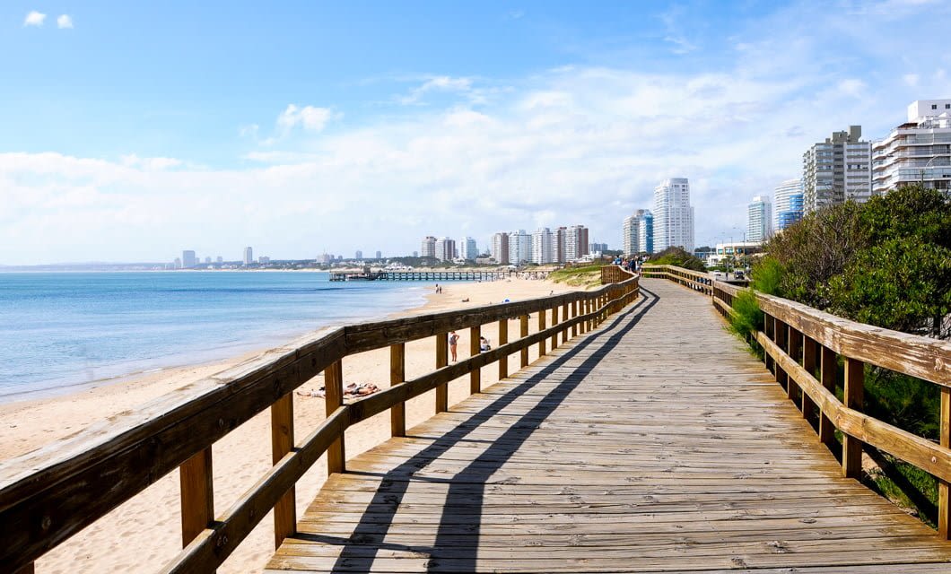 A wooden boardwalk runs along a sandy beach with blue water and green bushes in Uruguay. Tall modern buildings line the coastline of Montevideo in the distance under a partly cloudy sky.