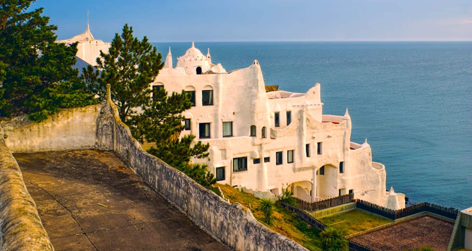 White, Gaudí-inspired Casapueblo building with curved walls and towers overlooks the blue sea in Punta Ballena, Uruguay—a stunning stop on any coast tour. A tree and stone path are in the foreground under a clear sky.