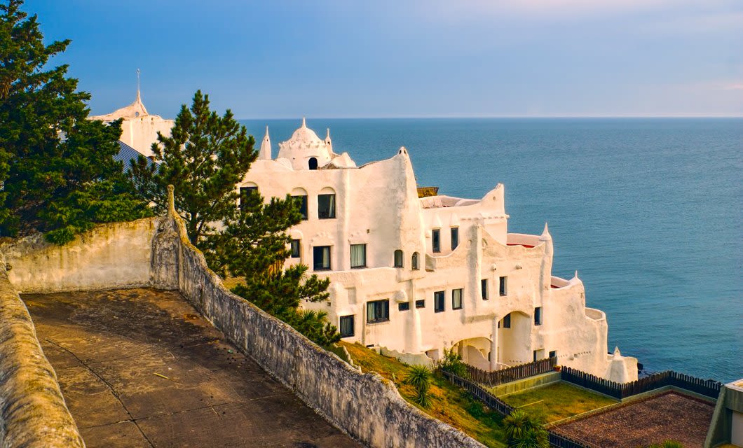 A white, whimsical building with organic shapes and multiple windows sits on a cliffside overlooking the ocean near Punta del Este, Uruguay, surrounded by greenery under a partly cloudy sky.