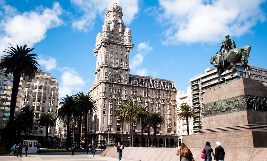 People walk in a city square in Uruguay with palm trees, a large ornate building with a tall tower, and an equestrian statue on a stone pedestal under a blue sky with scattered clouds.
