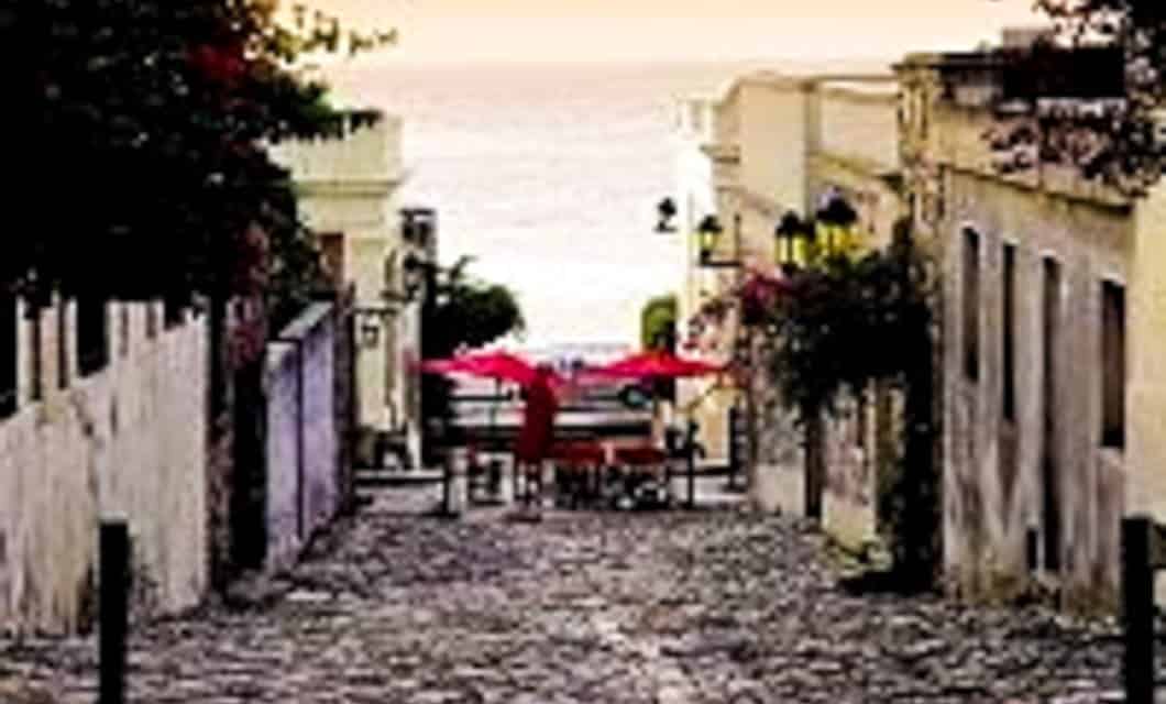 A cobblestone street slopes down toward a waterfront, lined with old buildings. At the end, red umbrellas cover outdoor tables where Tango and Samba rhythms drift through the air, with the sea visible in the background under a pale sky.