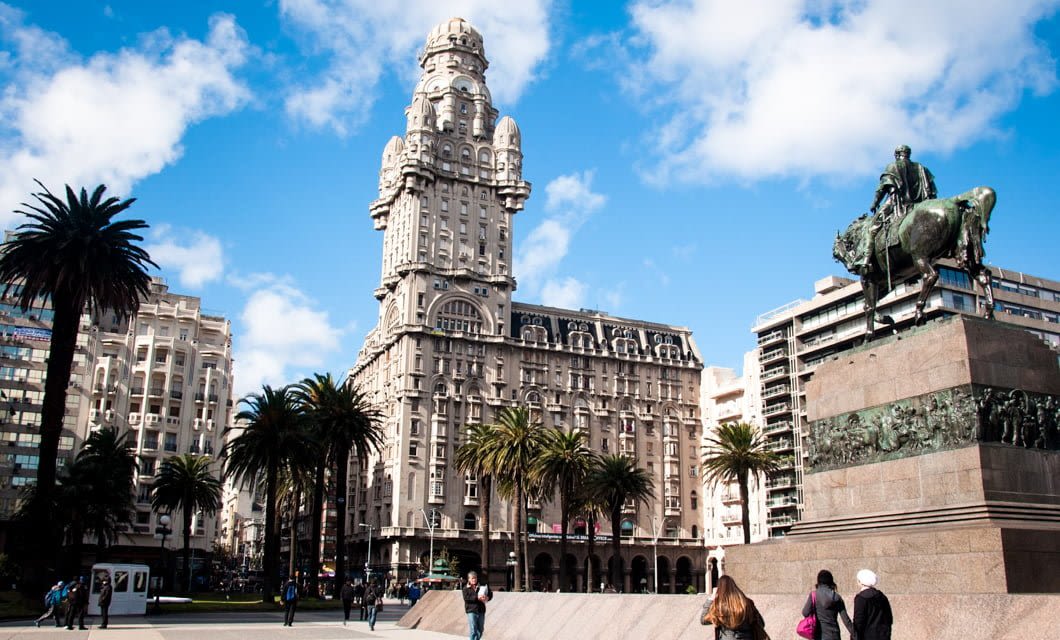 Plaza Independencia in Montevideo, Uruguay, features the historic Palacio Salvo, palm trees, people walking in rhythm with city life, and an equestrian statue beneath a bright blue sky dotted with clouds.