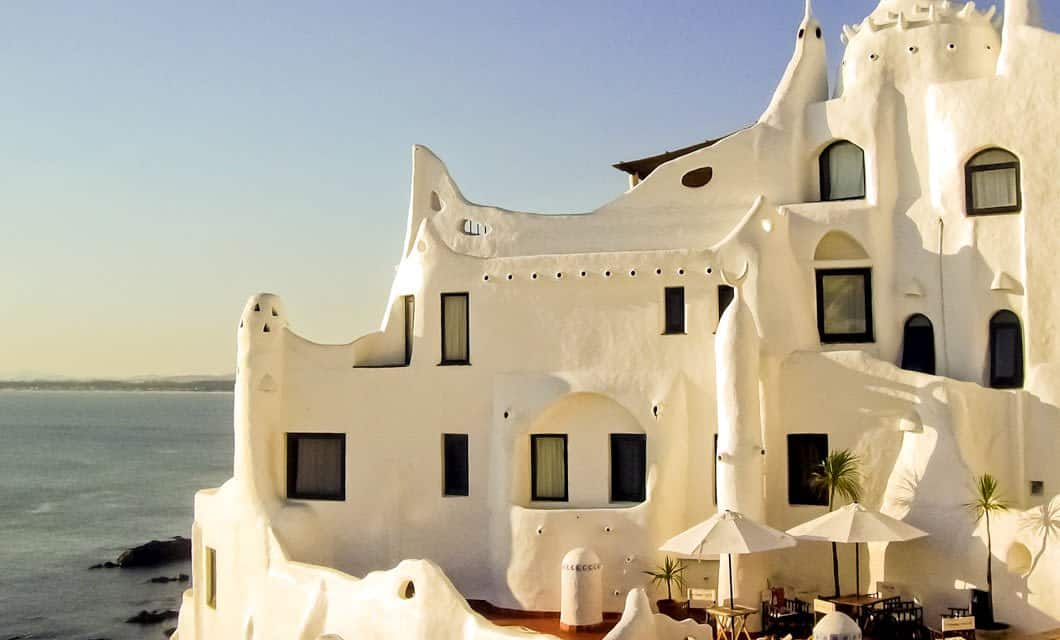 White, whimsical building with curved shapes and black-framed windows overlooks the calm sea of Uruguay. Patio tables with umbrellas and potted plants in the foreground rest under a clear sky, capturing the relaxed rhythm of coastal living.