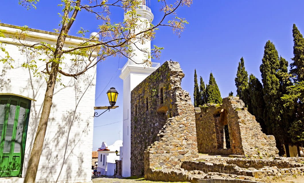 A tall white lighthouse stands next to old stone ruins and a white building with green shutters, framed by cypress trees and a clear blue sky—a hidden gem along your South America Golf Tour.