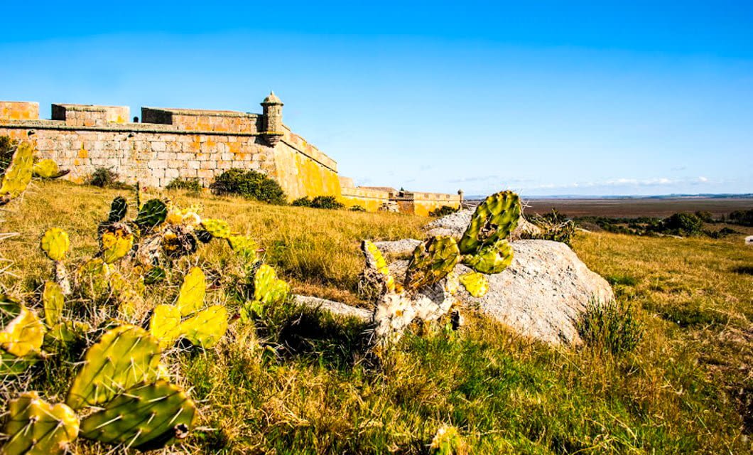 A historic stone fort stands on a grassy hill under a clear blue sky in Uruguay, with cacti and rocks in the foreground—a perfect starting point for horseback riding across the wide plain stretching into the distance.