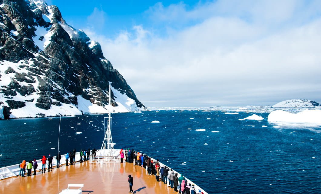 A group of people stand on the deck of an Australis Cruises ship, viewing snowy mountains and floating icebergs in the blue, icy sea of the Fuegian Fjords under a partly cloudy Patagonian sky.