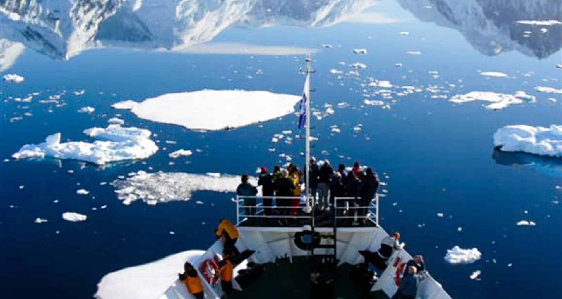 A group of people stand on the bow of a cruise ship navigating icy Antarctic waters near Ushuaia, surrounded by floating ice and snow-capped mountains, reflecting off the calm, blue water under a clear sky.