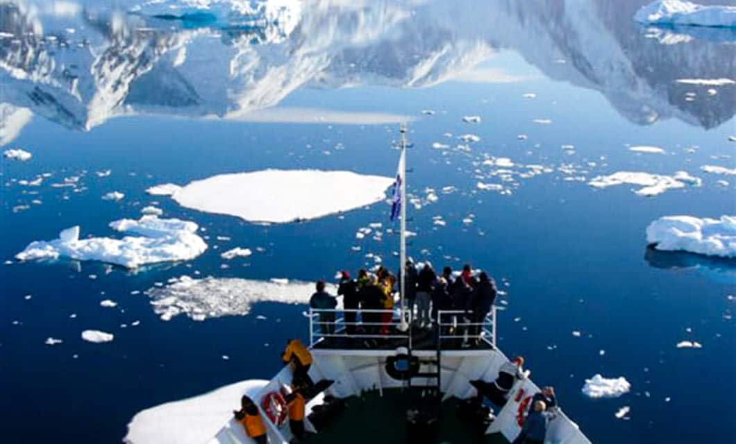A group of people stand on the bow of a ship during an Antarctica cruise, observing floating icebergs and calm blue water with snow-covered mountains reflected in the background.