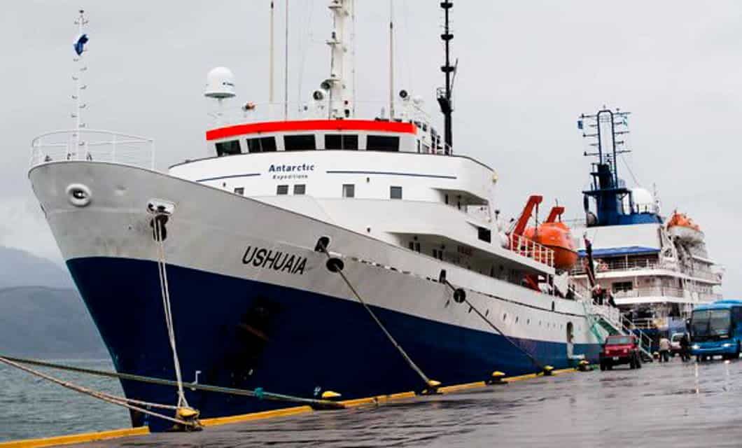 A large white and blue ship named USHUAIA is docked at a pier on a cloudy day. Another similar vessel and a blue bus are in the background. The ship’s sign reads Antarctic Expedition, hinting at a memorable Ushuaia cruise adventure.