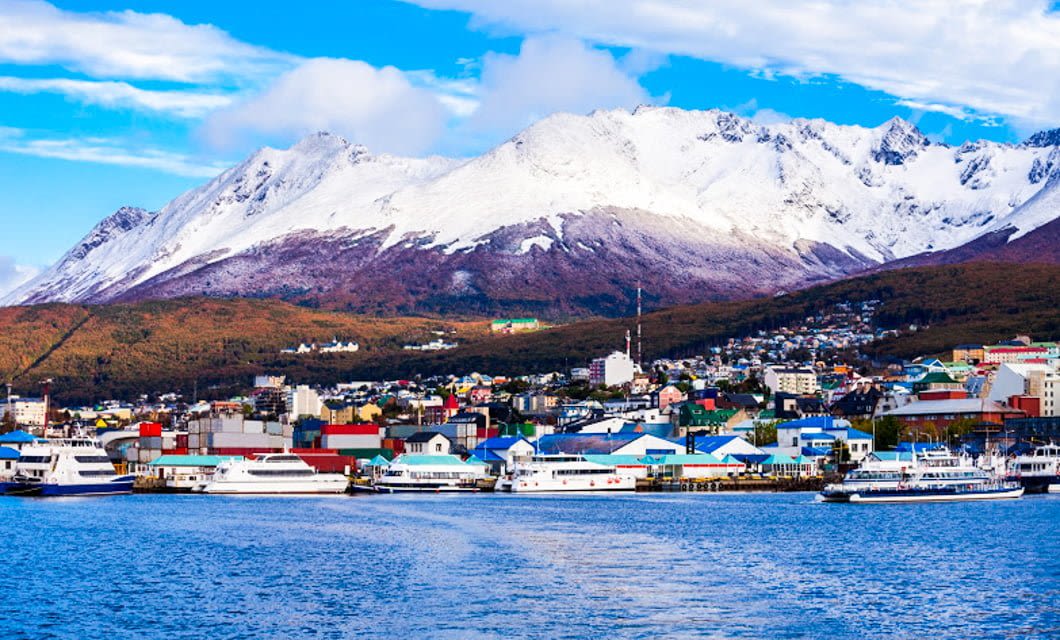 A coastal town with colorful buildings sits by the water in Ushuaia, with snow-capped mountains in the background and several boats, including the Plancius Antarctica cruise ship, docked along the shore under a partly cloudy sky.