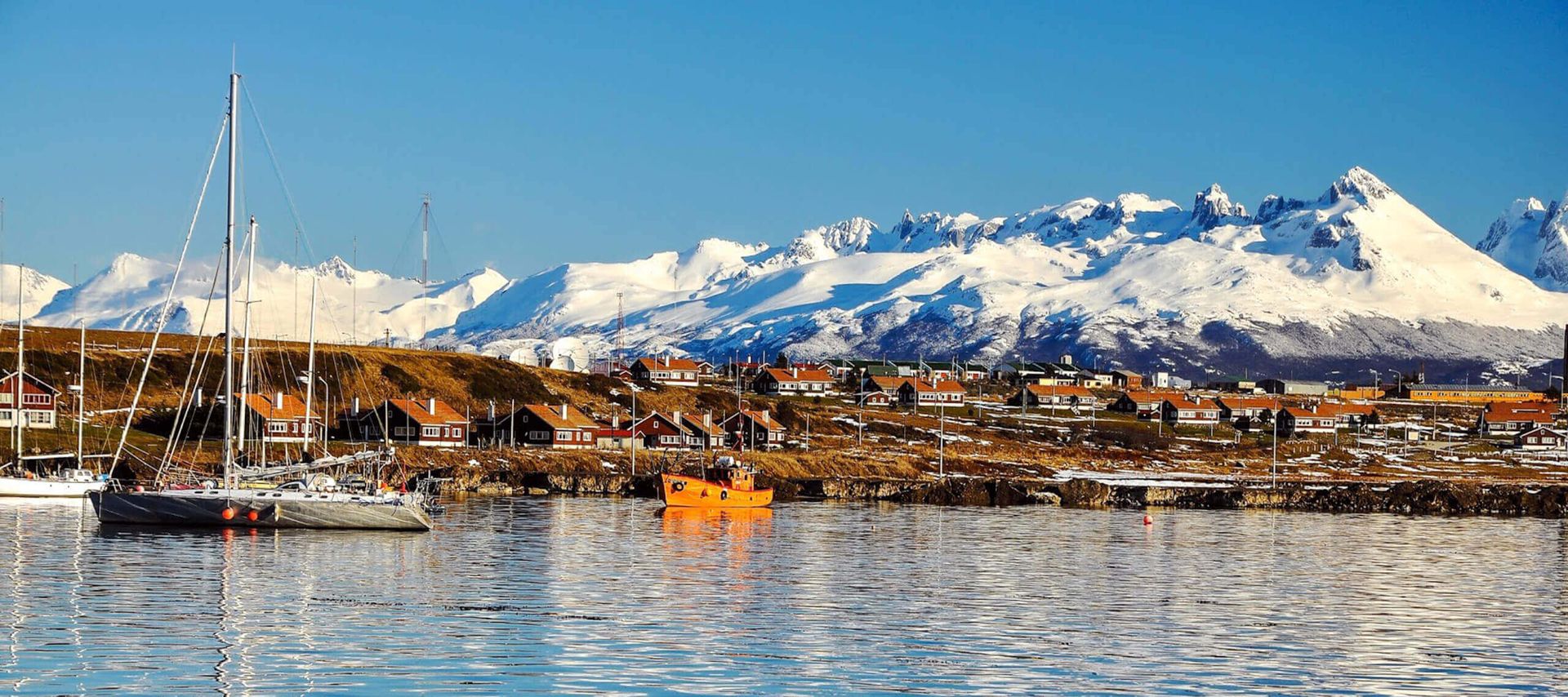Snow-capped mountains rise behind a coastal village with red-roofed houses. Sailboats and a bright orange boat float on calm, reflective water under a clear blue sky—an inviting scene for True Explorers in Argentina.