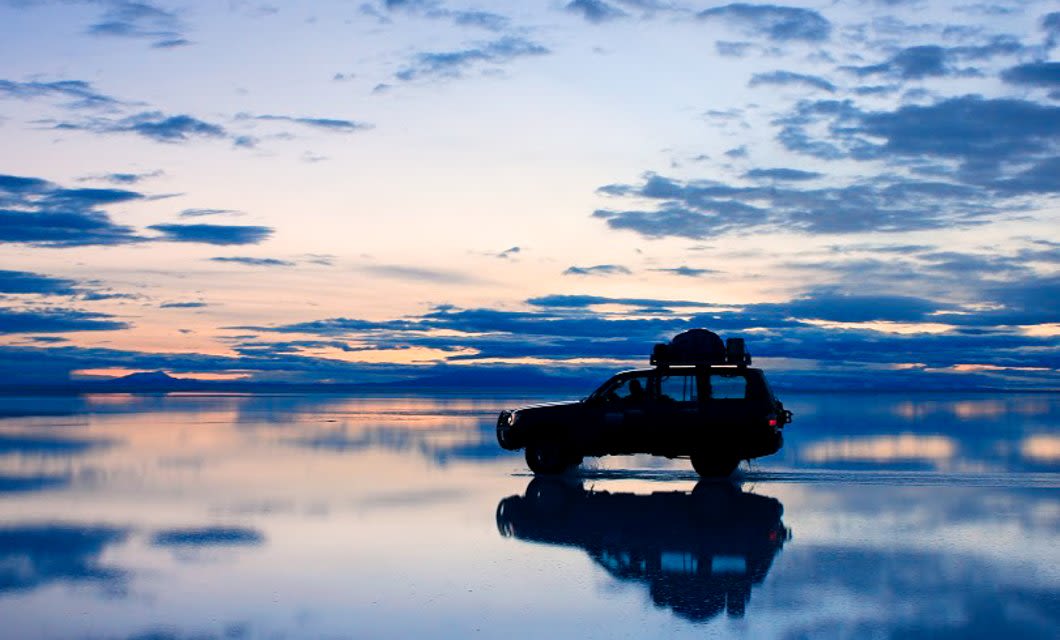 A silhouette of an SUV cruises across a reflective, water-covered surface under a blue and cloudy sunset sky—a striking scene that captures the breathtaking beauty of Bolivia highlights.