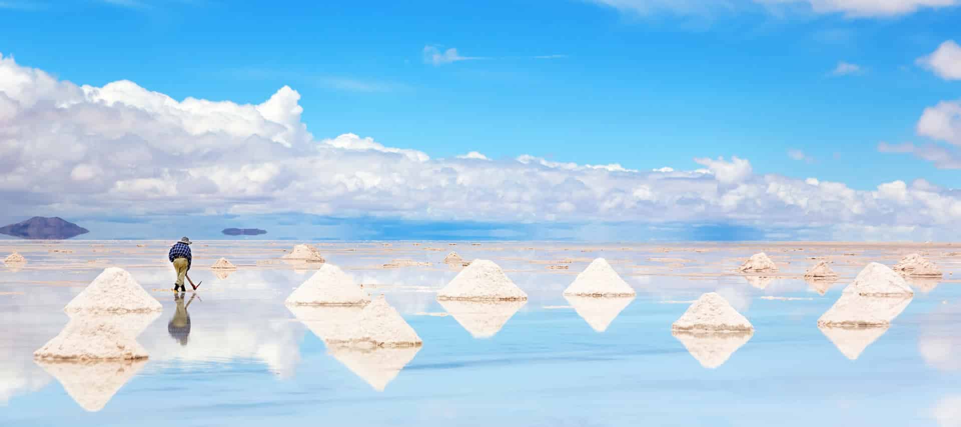 A person gathers salt on the stunning salt flats under a bright blue sky with clouds. Small white salt mounds are neatly arranged, creating mirror-like reflections reminiscent of scenes on Salar de Uyuni during Uyuni tours.