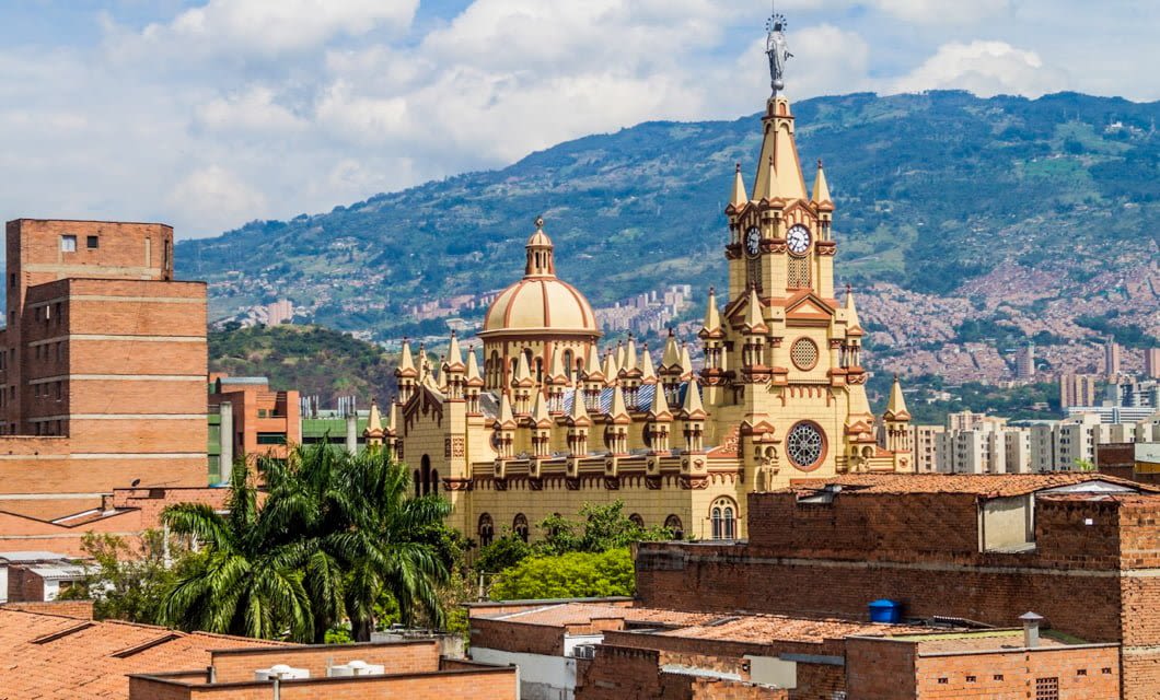 A historic yellow cathedral with a clock tower stands among red brick buildings in Colombia, set against lush green mountains and a blue sky—perfect for a scenic Botanical Tour.