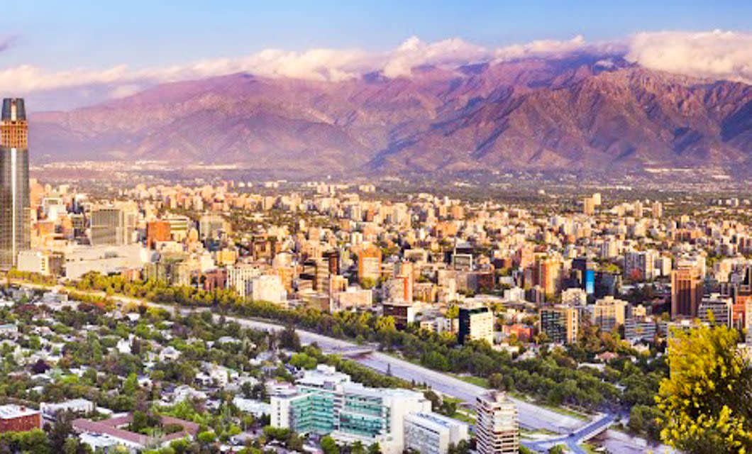 Cityscape of Santiago with modern buildings and skyscrapers set against tall, rugged mountains under a partly cloudy sky, with trees and a main road in the foreground—hinting at the proximity to the dramatic Atacama Desert.