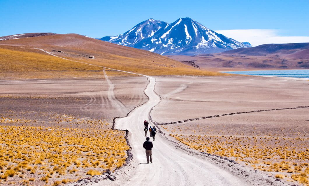A group of people walk along a winding dirt road through a dry, desert landscape in the Andes, with yellow grass patches and distant mountains capped with snow under a clear blue sky.