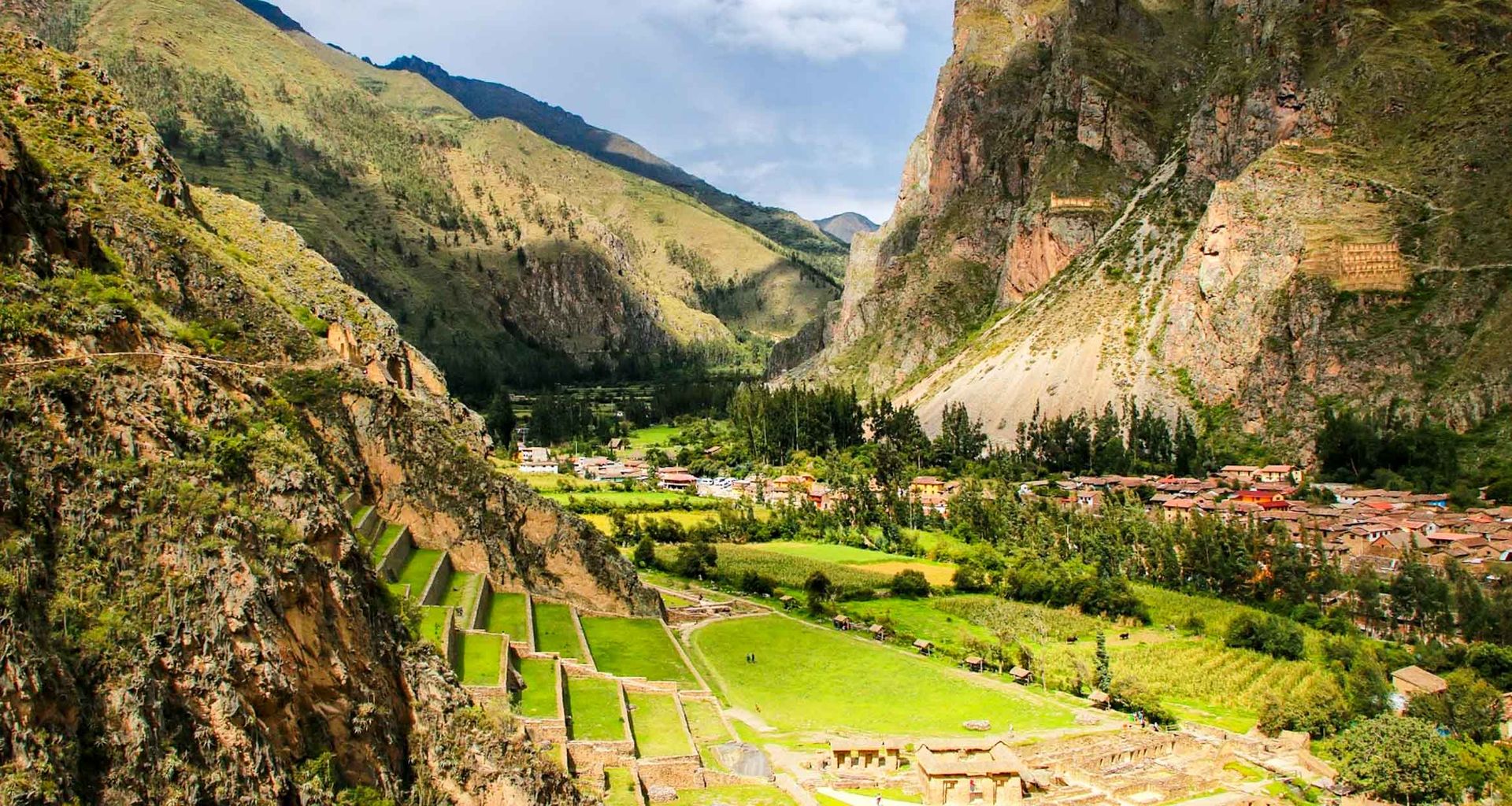 Terraced ancient ruins sit on a lush green mountainside along the Lares Trek, overlooking a valley with trees, fields, and a small village surrounded by steep rocky peaks under a partly cloudy sky.