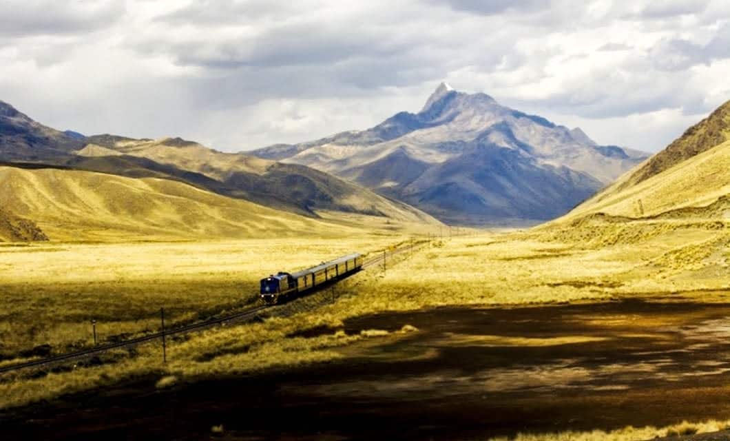 A blue train travels through a vast yellow grassland, once roamed by Gauchos, surrounded by rolling hills and distant mountains under a partly cloudy sky.