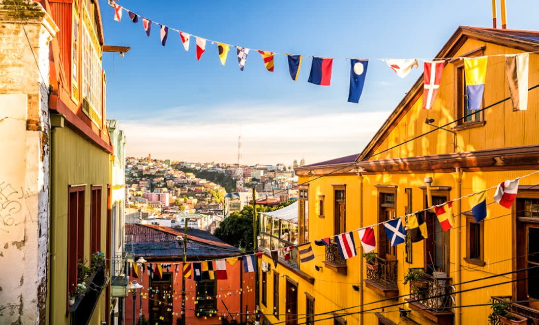 Colorful flags hang across a narrow street lined with vibrant yellow and orange buildings in Santiago, with a hillside cityscape and blue sky in the background—inviting you to explore more of this lively city.