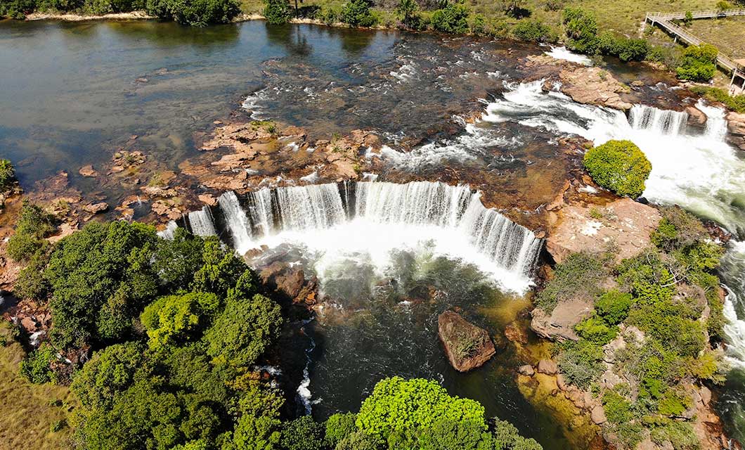 Aerial view of a wide, curved waterfall cascading over rocky ledges in Jalapão, Brazil's Cerrado, surrounded by lush green trees and clear, shallow water—an inviting sight for any tour.