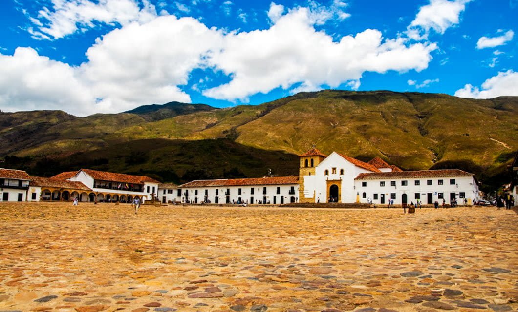 A wide view of a large cobblestone plaza in Boyacá, Colombia, with colonial-style white buildings and red-tiled roofs, set against green hills and a partly cloudy blue sky—a perfect spot for a memorable tour.