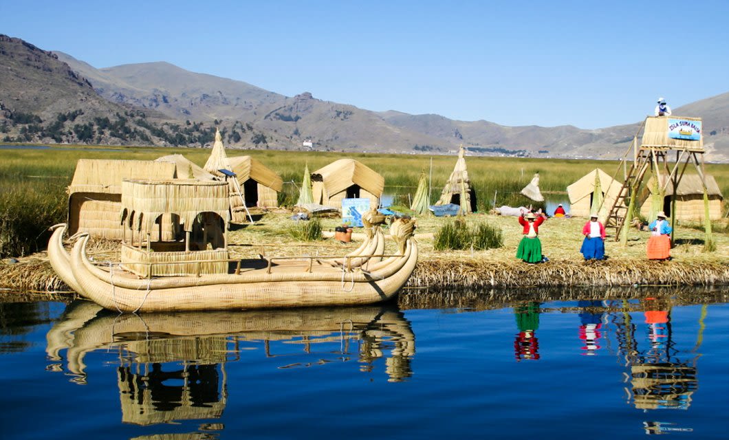 Traditional reed houses and a large reed boat sit on a floating island in Lake Titicaca, Peru. People in colorful clothing are seen on the island—an unforgettable stop on your Coast to Coast Tour—with mountains and blue sky in the background.