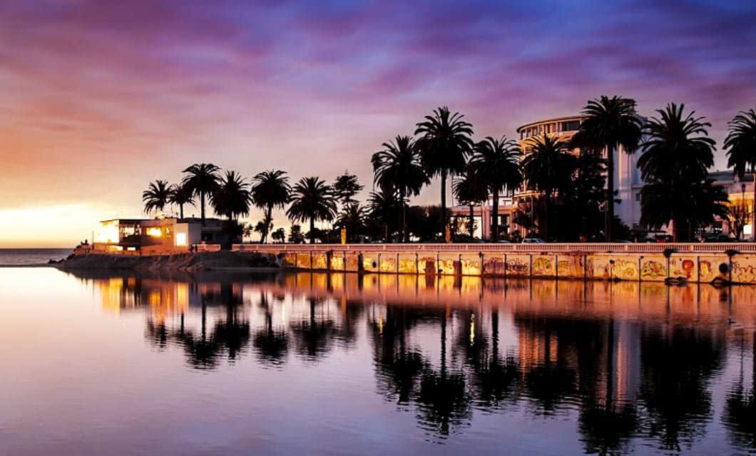 Palm trees and buildings in Santiago are silhouetted against a colorful sunset sky, reflected in calm water. Warm lights from the buildings and street lamps glow along the waterfront, creating an inviting scene perfect for an evening tour.