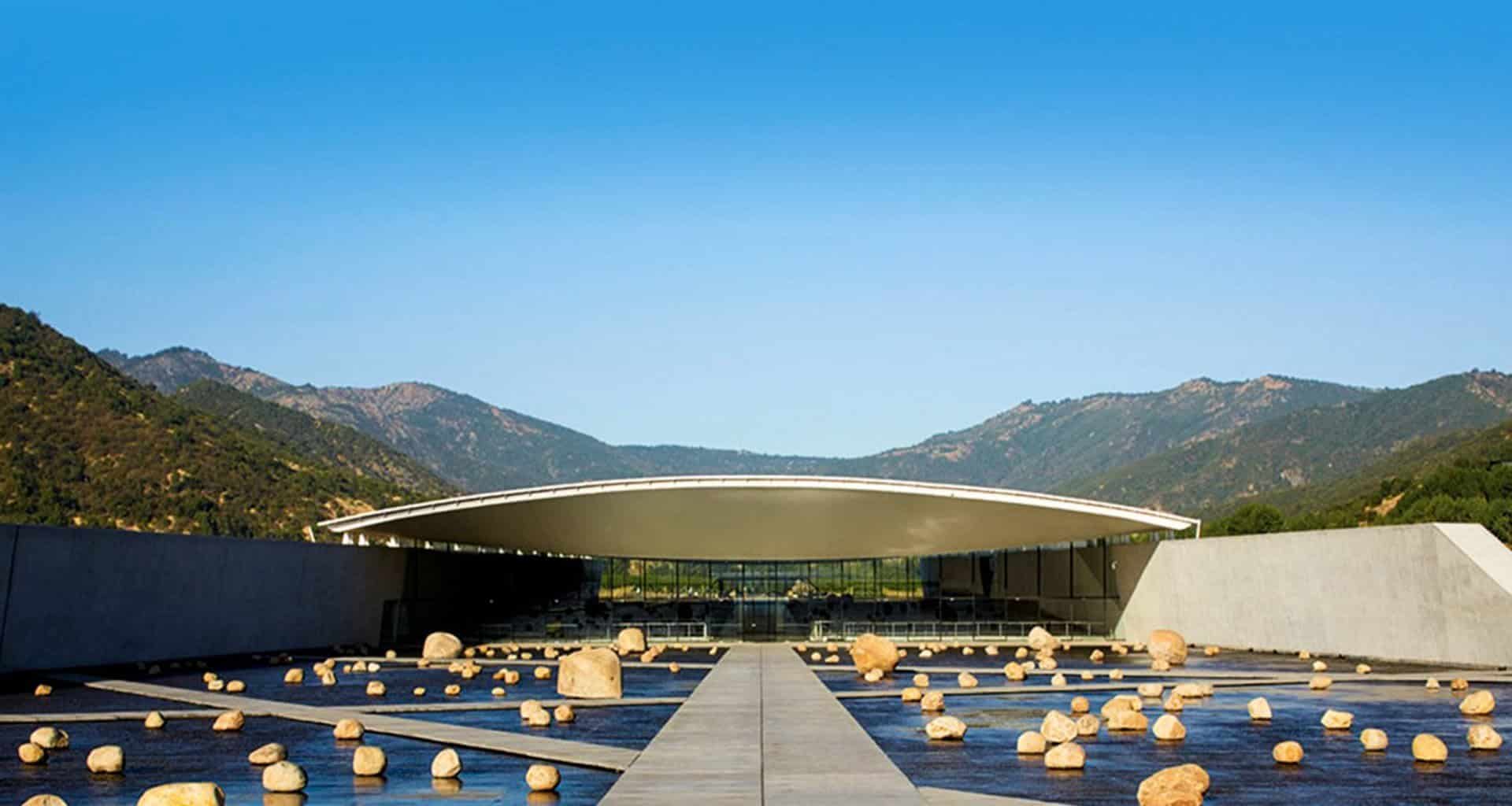 A modern building with a curved white roof, surrounded by mountains, is home to Viña Vik Hotel, a luxury hotel in Chile. A reflecting pool with scattered large rocks completes the serene scene beneath the clear blue sky.
