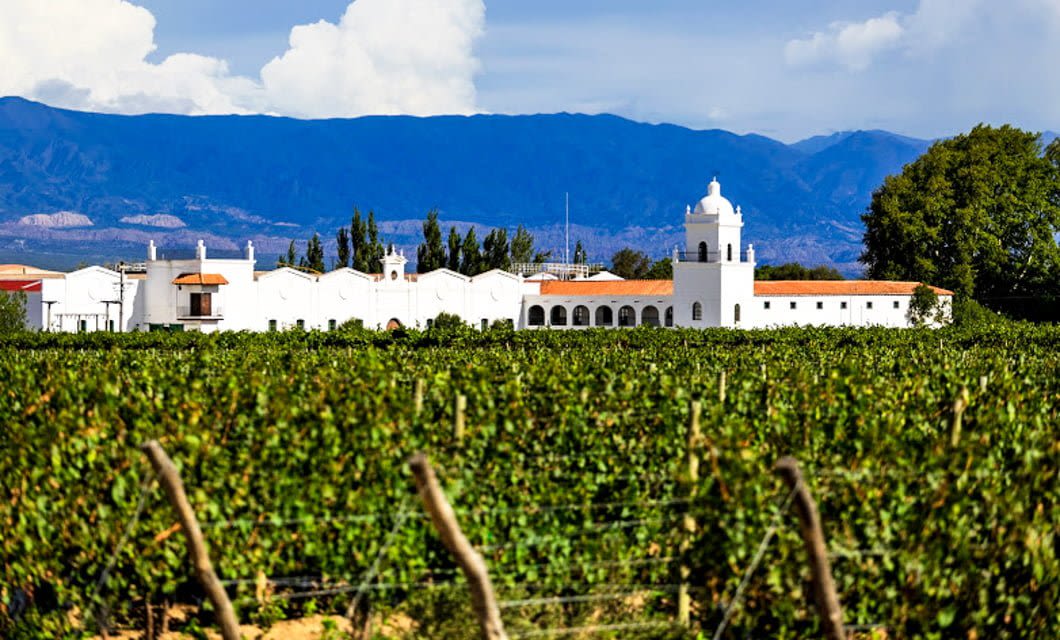 A white colonial-style building with a tower sits behind lush green vineyards at one of the renowned wineries of Argentina, framed by tall trees and mountains under a partly cloudy blue sky.