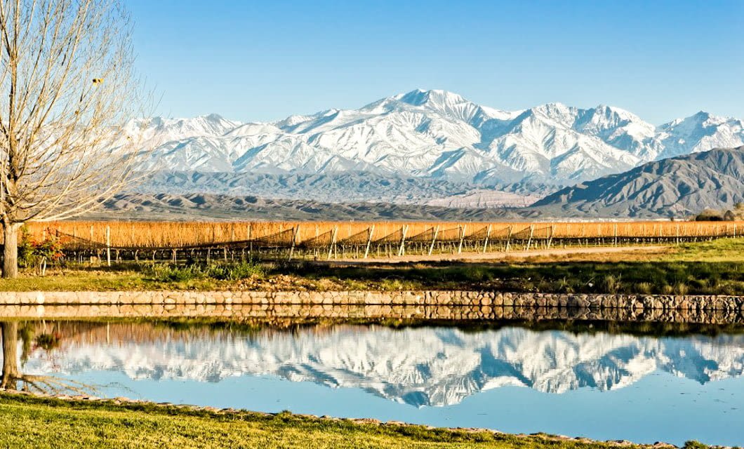 A tranquil pond reflects snow-capped mountains and a clear blue sky, with golden fields and bare trees in the foreground—an idyllic South America scene perfect for a serene wine tour.