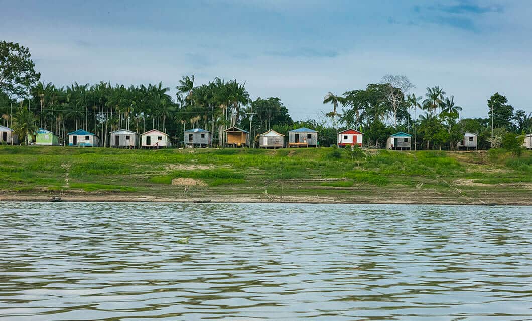 A row of small, colorful houses stands on a grassy riverbank in the Remote Amazon, with tall palm trees behind them under a blue sky, as seen from across the rippling water.