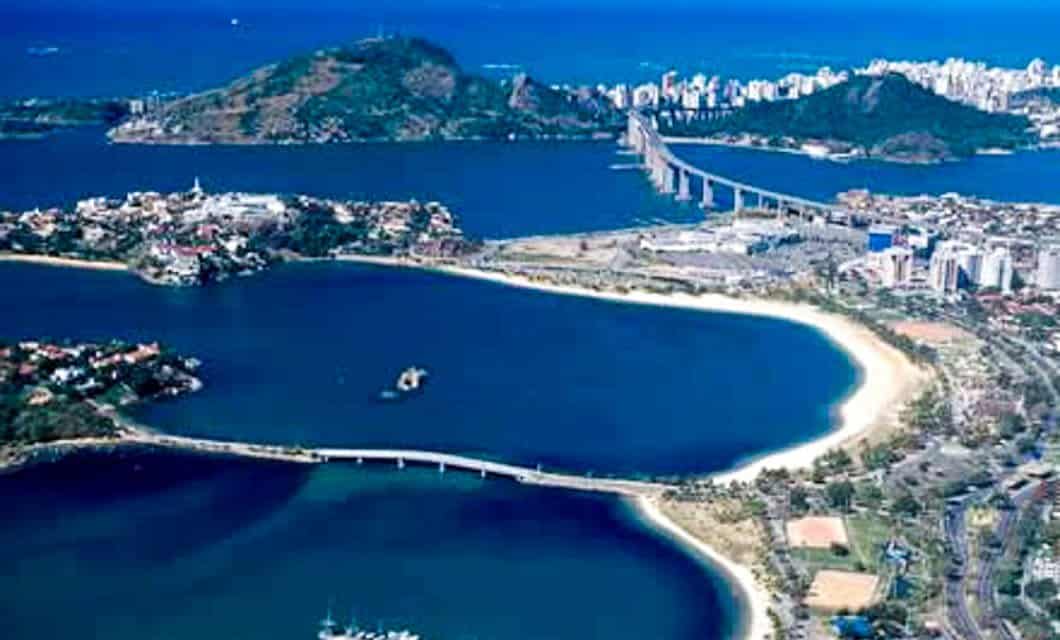 Aerial view of a coastal city with sandy beaches, blue water, bridges connecting urban areas, and green hills in the background. Explore vibrant shorelines and buildings as part of a Brazil Train Tour along the scenic Brazil Gold Route.
