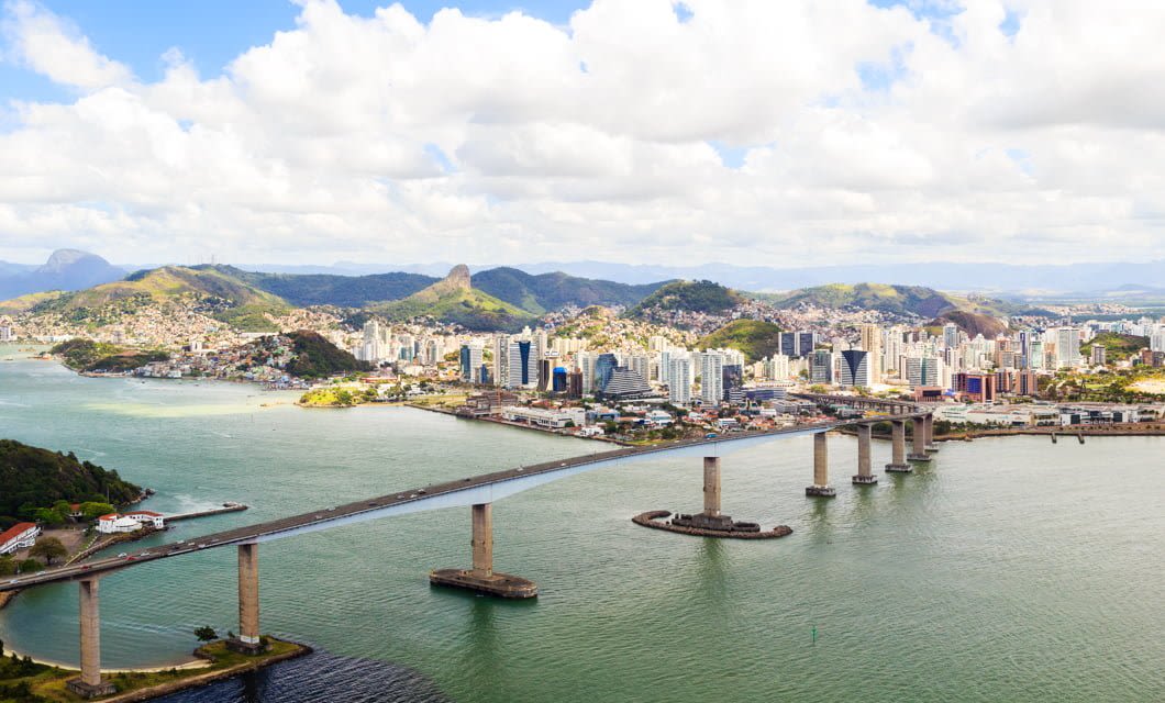 A large bridge crosses over water toward a city with many tall buildings, surrounded by green hills and mountains under a partly cloudy sky—an ideal view for those experiencing a Brazil Train Tour.