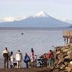 People stand on a rocky lakeshore, looking out at the water. In the background, a snow-capped volcano rises above the horizon under a partly cloudy sky. A wooden pier extends into the lake on the right, showcasing one of nature’s stunning lakes.
