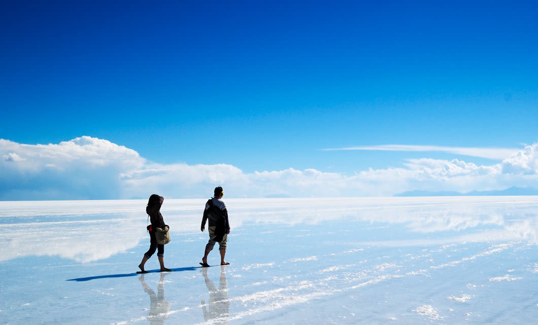 Two people walk across a reflective, water-covered salt flat under a bright blue sky with scattered clouds, creating the illusion they are walking on the sky—an unforgettable sight on a Bolivia train tour.