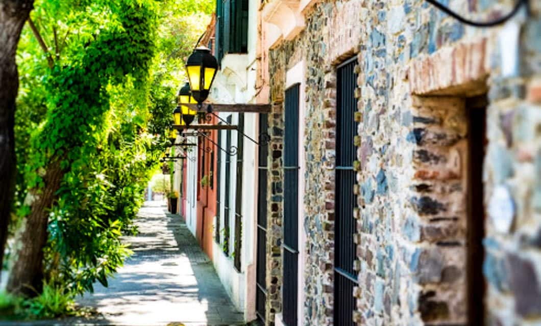 A narrow cobblestone street lined with stone buildings, black iron lamps, and lush green trees casting dappled sunlight on the sidewalk—echoes of tango music drifting softly through the afternoon air.